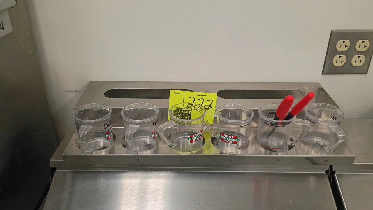 Stainless steel measuring cup holder. Features cutout inserts for portion cups and rear storage slots. Includes (6) plastic measuring cups. 23" x 10".