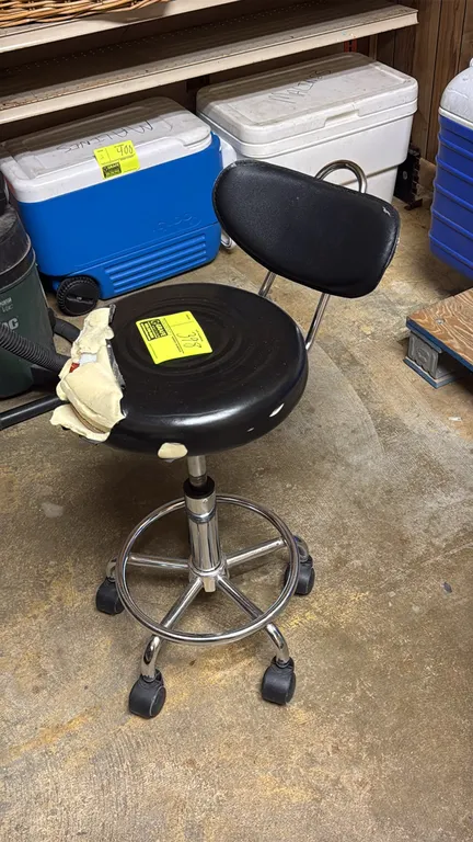 Rolling swivel stool. Features black vinyl upholstered seat and backrest, integrated foot ring, and pneumatic height adjustment. Mounted on a chrome 5