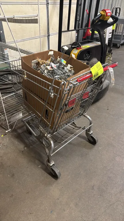 Wire shopping cart with an assortment of display peg hooks. Features metal wire basket construction, red push handle, lower tray, and a cardboard box