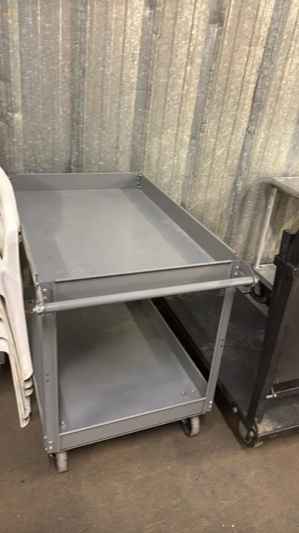 Metal 2-tier utility cart. Features two lipped tray shelves and an integrated push handle. 36"L. Mounted on casters.