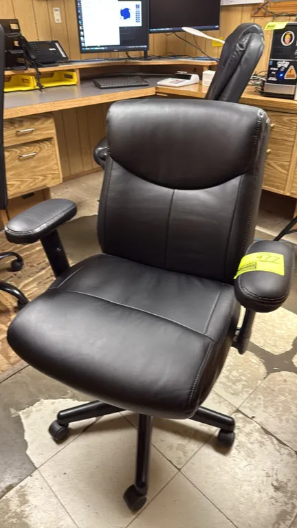 Executive office chair. Features black faux leather upholstery, padded armrests, and cushioned seat and back. Mounted on a 5-star base with rolling ca
