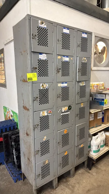 18-compartment metal employee locker unit. Features 3-column, 6-tier configuration, perforated ventilation doors, and padlock hasp latches. Gray paint