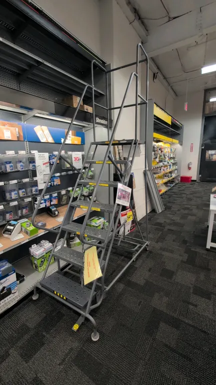Rolling metal stock ladder. Features 7-step construction with perforated slip-resistant treads, tubular steel frame, dual side handrails, and upper gu