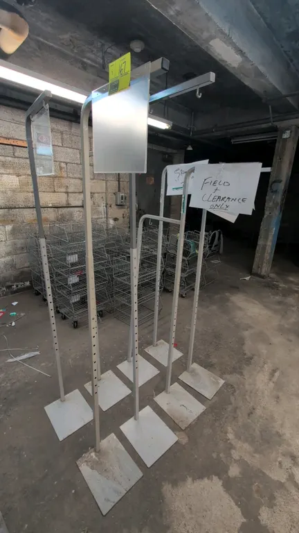 Adjustable metal sign stands. Features height-adjustable upright posts with hanging hooks and flat square metal floor bases. Includes clear plastic si
