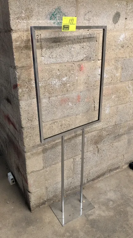 Metal floor sign stand. Features a chrome-finish frame with slide-in sign channel and dual upright support posts. Mounted on a flat rectangular base.