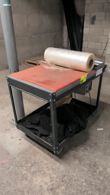 Portable metal shrink wrap station. Features dual film rollers, top work surface, integrated power control box with auxiliary outlet, and lower storag