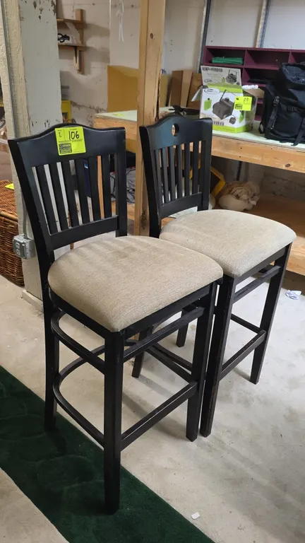 Wood frame bar stools. Features beige upholstered cushioned seats, vertical slat backs with circular cutout handle accents, and integrated footrest st