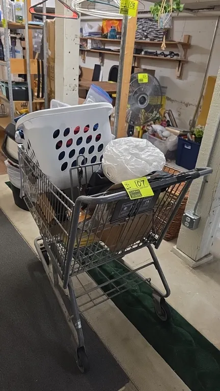Metal grocery cart with assorted contents. Includes full-size metal wire shopping cart, plastic laundry basket, assorted fabric, clothing, linens, and