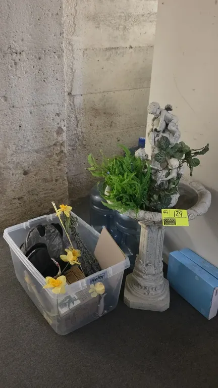 Resin cherub pedestal fountain and Hefty plastic storage bin with assorted contents. Fountain features a tiered cherub motif with faux greenery accent