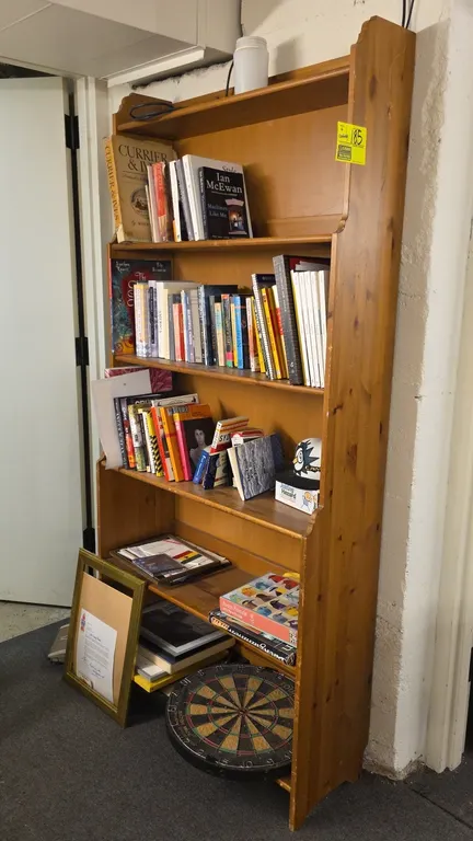 Wood 6-tier shelving unit with assorted contents. Features a shaped side profile and natural wood finish. Includes assorted books, board games, framed