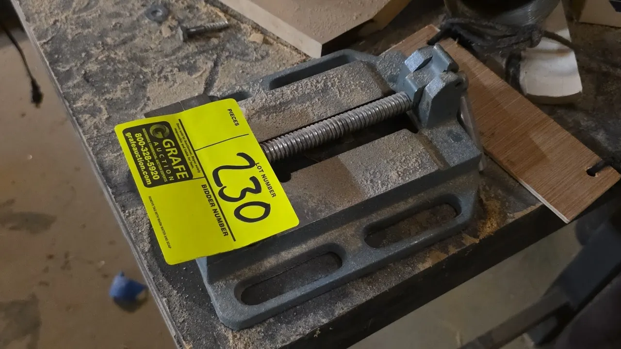 Metal drill press vise table clamp. Features cast metal construction, threaded adjustment screw with lever handle, and slotted mounting base flanges.