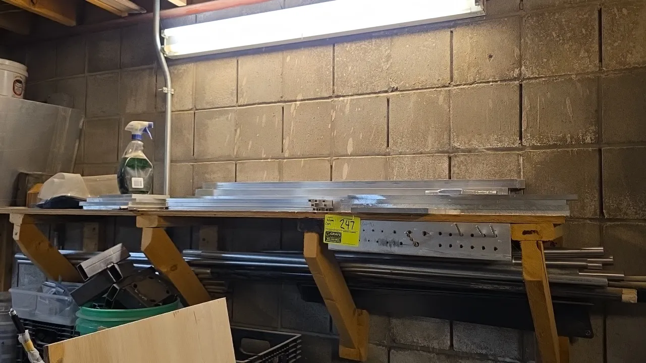 Assorted aluminum raw material stock. Includes various aluminum flat bar, plate stock, extrusions, and round tubing. Note: Wooden shelving, drill bits