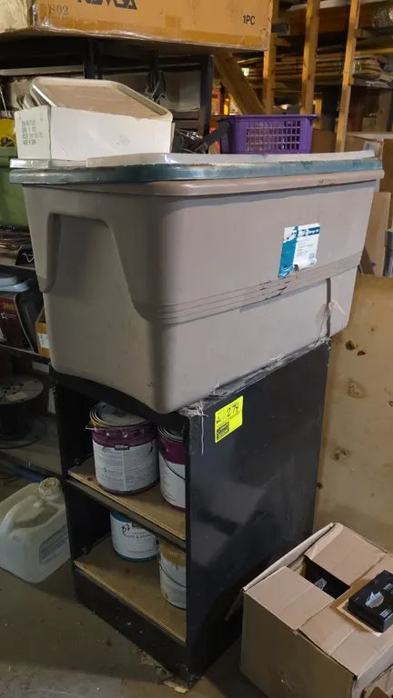 Metal 2-tier shelf unit with plastic storage tote and lid. Features black metal construction with interior shelf inserts. Contents not included.