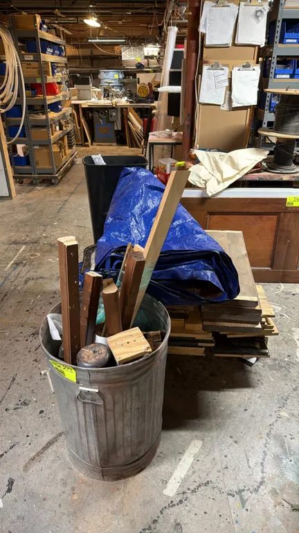 Assorted scrap wood, tarps, and trash cans. Package lot includes a galvanized metal trash can with assorted wood pieces, a black plastic wastebasket,