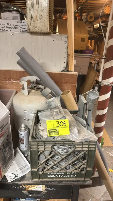 Barbed wire coil and fencing hardware. Includes galvanized barbed wire and fence fittings housed in a plastic milk crate. Propane tank and background