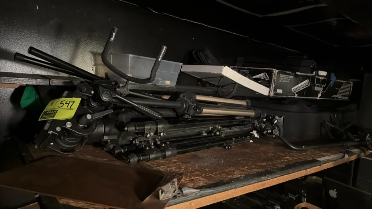 Assorted camera, microphone, and equipment stands. Features folding tripod bases and adjustable legs. Shelving, boxes, and surrounding background item