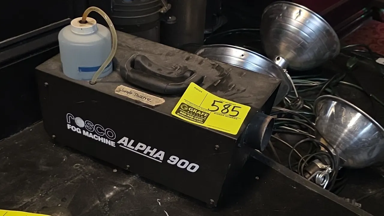 Rosco Alpha 900 fog machine. Features a top carrying handle, integrated fluid reservoir with feed tube, and black metal housing. Background items not
