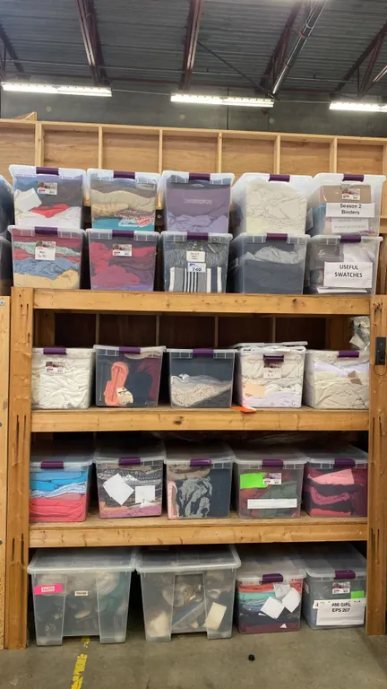 GROUP OF 25 PLASTIC STORAGE BINS FULL OF ASSORTED FABRIC MATERIAL AND 4 ROLLS OF FABRIC