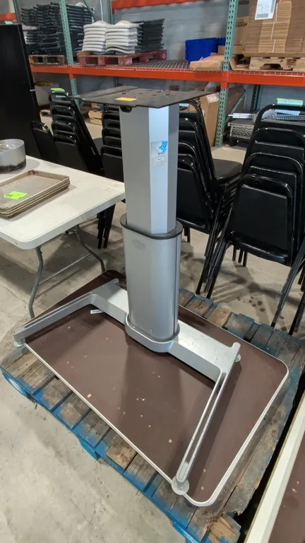 Pneumatic sit-stand desk base. Features a single pneumatic height-adjustable telescoping column with an upper mounting plate and a silver-tone metal b