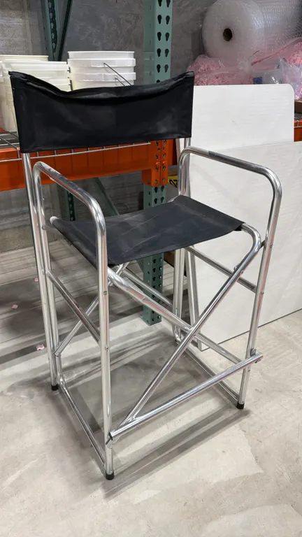 Folding director's chair. Features black fabric seat and backrest, integrated armrests, and folding footrest. Mounted on an aluminum frame.