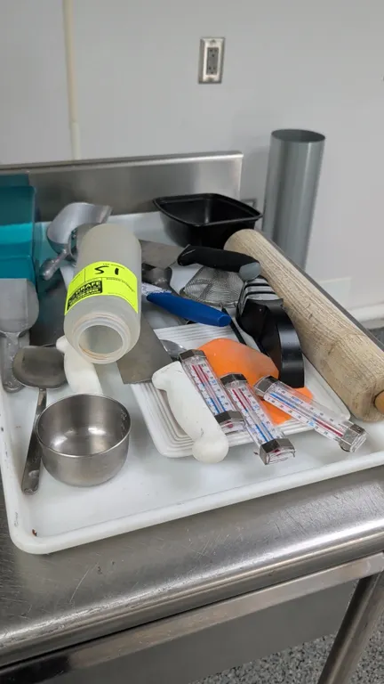 Full-size poly bakery tray with assorted kitchen utensils. Includes a wooden rolling pin, thermometers, kitchen knives, handheld knife sharpener, meta
