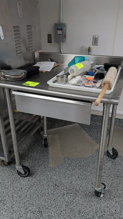 Stainless steel work table. Features a rear backsplash and an under-counter sliding drawer. 36" x 30". Mounted on casters. Contents not included.