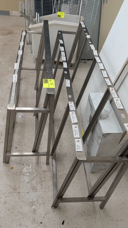 Stainless steel angled tray display racks. Features all-welded tubular frame construction and shelf labeling channels. Dimensions: 56"W and 74"W.