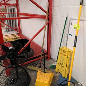 Assorted maintenance and cleaning supplies. Includes Lasko electric pedestal fan, push brooms, angle broom, upright lobby dustpan, hand dustpan, and f
