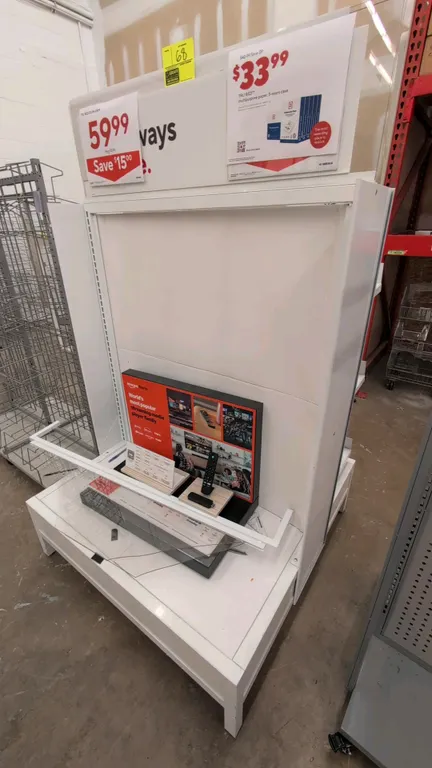 Double-sided metal merchandise display rack. Features white finish, three sloped tiered shelves with front retaining lips on one side, flat base deck