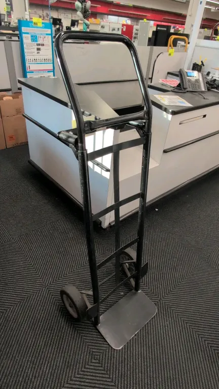 Metal hand truck. Features black painted tubular steel construction, loop handle, straight back crossbars, and solid nose plate. Mounted on two solid