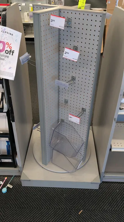 Rotating pegboard merchandise display rack. Features a 4-way rotating pegboard tower with grey laminate frame, assorted metal peg hooks, and clear acr