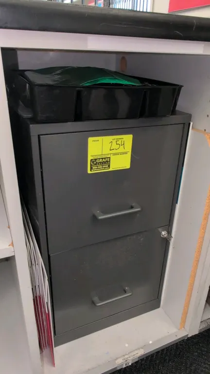 Metal 2-drawer vertical filing cabinet. Features a gray finish, integrated pull handles, and lock with key. 14"W. Contents and surrounding cabinetry n