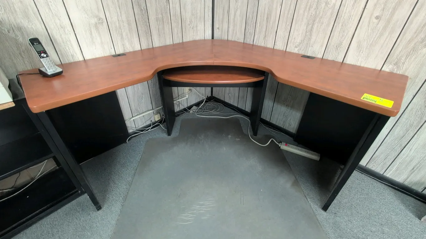 Laminate wood L-shaped corner desk. Features a pull-out keyboard tray, cherry-tone top finish, black panel base, and integrated wire grommets. 61" x 6