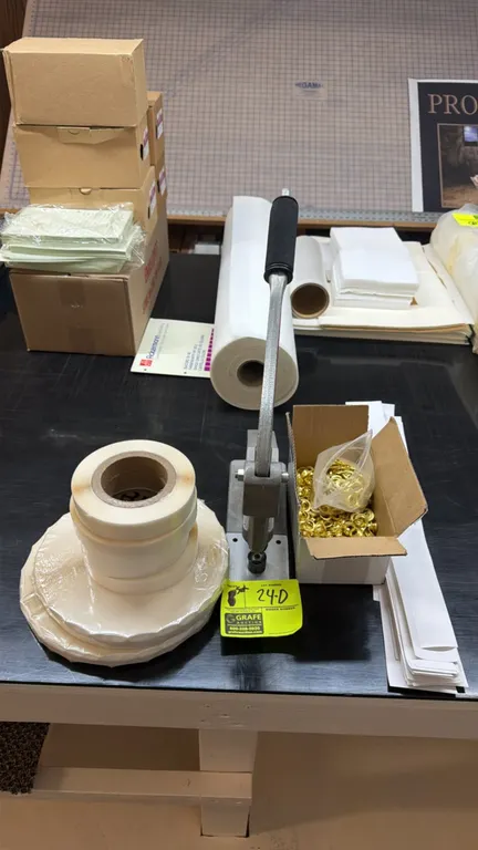 Manual grommet press and banner finishing supplies. Includes hand-lever brass ring press, box of brass grommets, rolls of double-sided tape, and paper