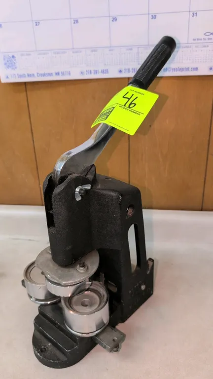 Press Master manual button maker press. Features cast metal frame, sliding dual die base, and manual lever handle with ribbed grip. Bench mountable.