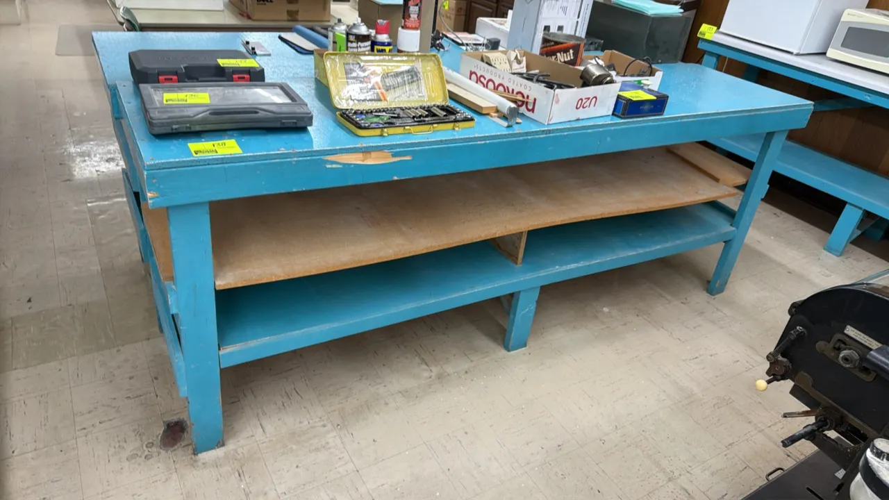 Wooden work table. Features blue painted wood construction with two lower storage shelves. 8'L x 2'W x 3'H. Contents not included.