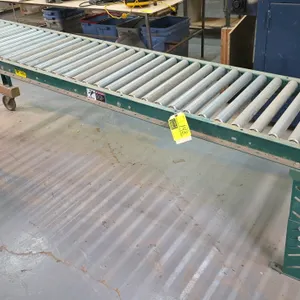 Gravity roller conveyor. Features galvanized steel rollers, green steel frame, wooden end stop, and adjustable-height legs with casters on one end. Di