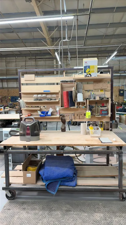 Portable work table. Features a wood top, wooden slat organizer backboard with assorted storage shelves and cubbies, and a lower wooden shelf. Mounted