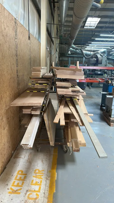 Assorted lumber and wood stock on a double-sided rolling material rack. Includes various wood boards, trim, sheet goods, and millwork pieces. Multi-ti