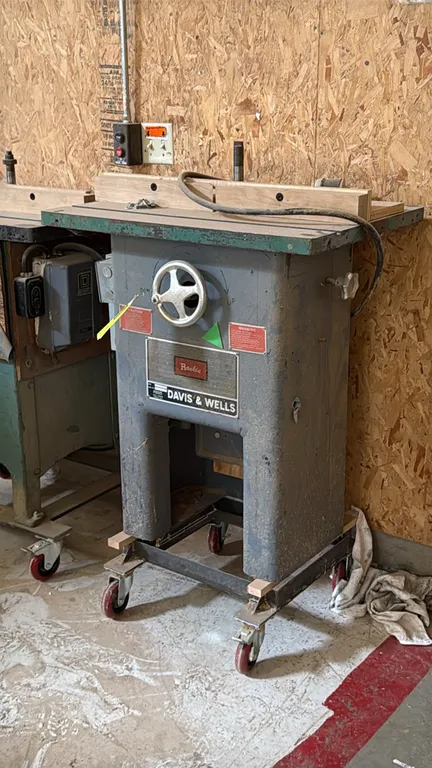 Davis & Wells wood shaper. Features a wooden fence assembly, cast iron table top, front spindle height handwheel, and attached electrical switch box.