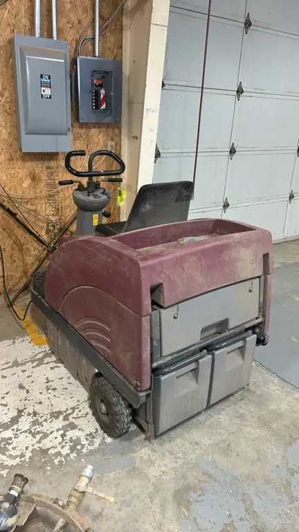 Minuteman Kleen Sweep ride-on floor sweeper. Features right-side curb broom, operator seat, steering column controls, and rear debris hoppers. Battery