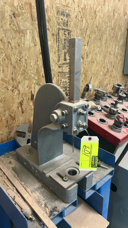 Manual arbor press. Features square toothed rack ram, base mounting plate with center cutout, and slotted anvil plate. Mounted on a blue welded steel