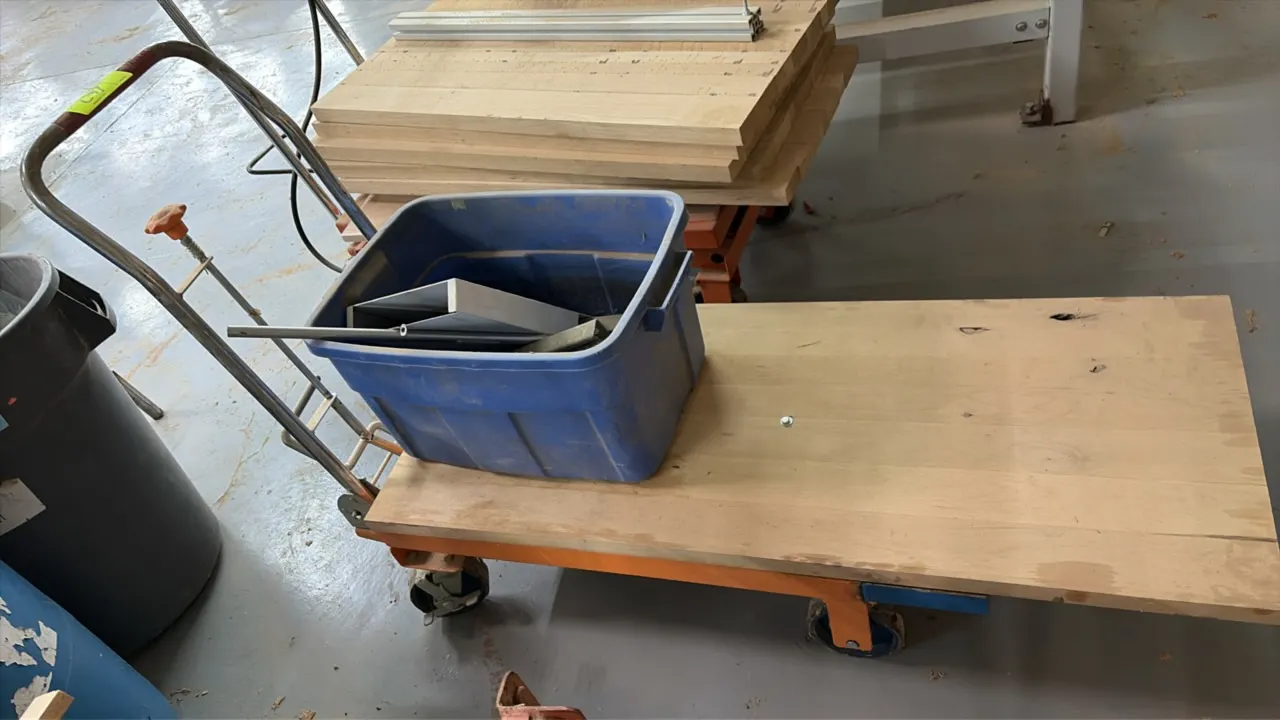 Hydraulic scissor lift table cart. Features foot pedal pump operation, manual lowering knob, extended wood top deck, and tubular steel push handle. Mo