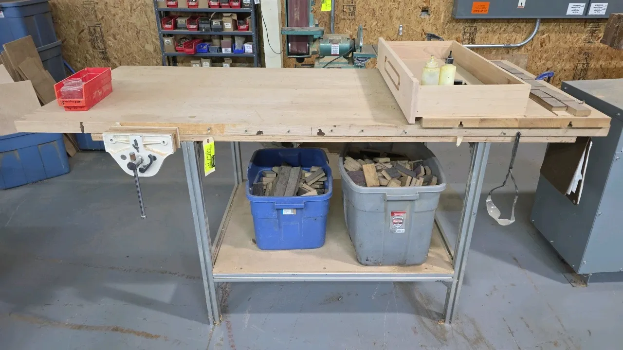 Wood top metal frame workbench. Features an integrated front woodworking vise, lower storage shelf, two plastic storage totes with wood cutoffs, and r