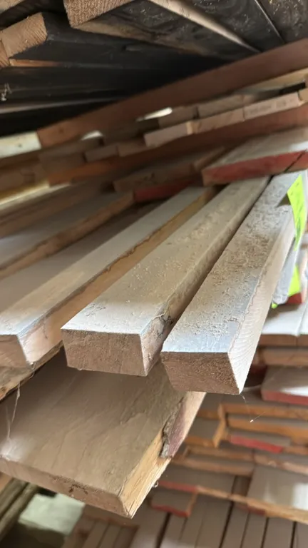 Skid of assorted wood boards. Features lumber of assorted widths and thicknesses. Approx. 10'L x 3'W x 1'H stack. Surrounding materials and racking no