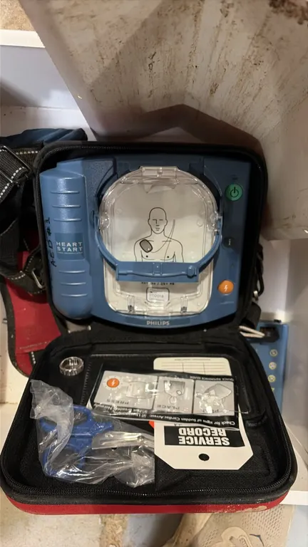 Philips HeartStart automated external defibrillator (AED). Includes red zippered carrying case, trauma shears, and a white metal wall-mount storage ca