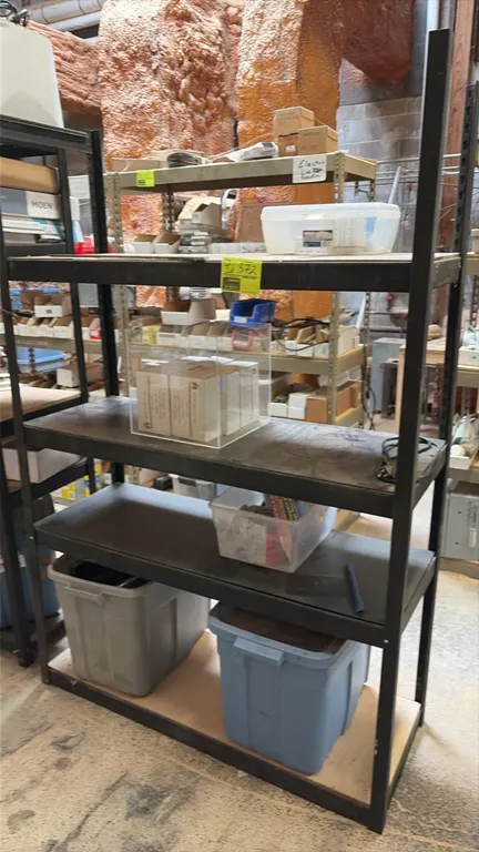 Metal storage rack with assorted parts and contents. Includes 5-tier black metal shelving unit, acrylic display case containing boxed staples, and pla
