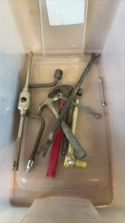 Assorted hand tools and drill bits with plastic storage tote. Includes axe with wood handle, large auger and core drill bits, hammers, pry bar, tap ha
