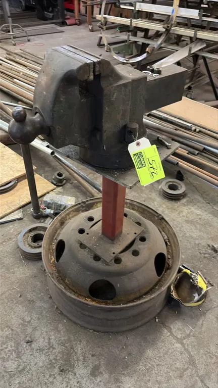 Rock Island 6" bench vise. Features cast iron construction with integrated anvil. Mounted on a heavy-duty steel pedestal stand with a wheel rim base.