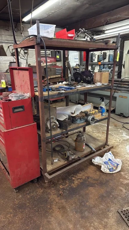 5' metal storage rack with contents. Features 4-tier steel frame construction with wood shelves. Includes lathe chucks, faceplate, rotary indexing tab